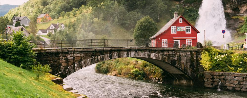 A bridge over rushing water. A red wooden house and a small mountain in the background.